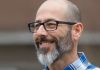 We Are the Virus Smiling man with glasses and a beard wearing a blue checkered shirt.