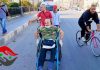 “I am carrying a message saying that our wounds are the key to our resilience and our victory.”/ Syria Insider A man in camouflage shirt sitting in a wheelchair pushed by another man in a red shirt outdoors.