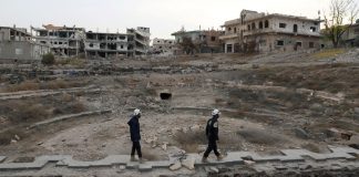 Decision to bring White Helmets to Canada dangerous and criminal/By Eva Bartlett, August 10, 2018/RT Two people in protective gear walk through a heavily damaged urban area.