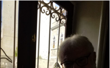 “Why sanctions? Why hurt the poor people?” Elderly man with glasses sitting by a window in dim light.