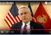 We Do Not Care How Many Ukrainians Die/ By Richard Hayden Black A man speaking in front of American and Marine flags.
