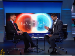 Two men in suits engaged in a televised interview on a modern set.