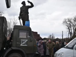 A statue with a blue bucket and people gathered around vehicles.