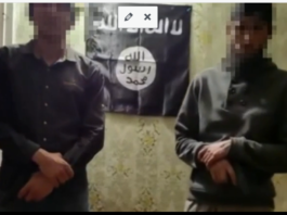 Two men standing in front of a black flag with Arabic script.