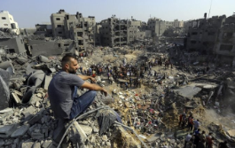 Man sitting amid extensive rubble in a heavily damaged urban area.