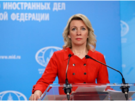 A woman in a red outfit speaking at a podium with a blue background.