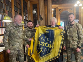 Four soldiers in camouflage holding a yellow and blue flag indoors.