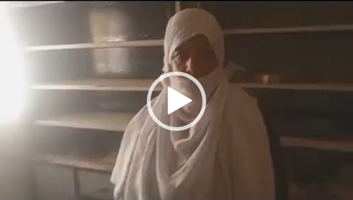 A person in white traditional attire stands indoors with shelves in the background.
